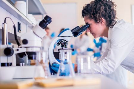 Une chercheuse est dans un laboratoire, penchée sur une table et regardant dans un microscope avec des tubes à essai tout autour d'elle.